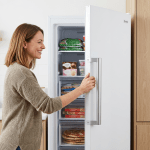 Woman opening a white freezer containing frozen vegetables, ice cream, frozen meals, and pizzas